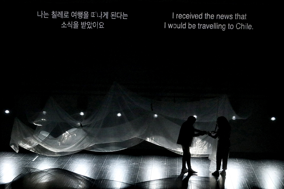 얇은 천이 겹겹으로 길게 드리워진 어두운 무대에서 힐다와 에바나가 손을 잡고 마주 서 있다. 상단 자막에는 ‘나는 칠레로 여행을 떠나게 된다는 소식을 받았어요’라는 말이 한국어와 영어로 쓰여 있다.