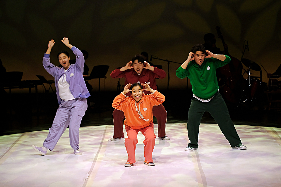 큰 원으로 비추는 조명 아래 다른 색깔의 트레이닝복을 입은 네 명의 출연자가 경쾌한 몸짓과 표정으로 팔을 크게 들어 올린 동작으로 함께 퍼포먼스를 펼치고 있다.