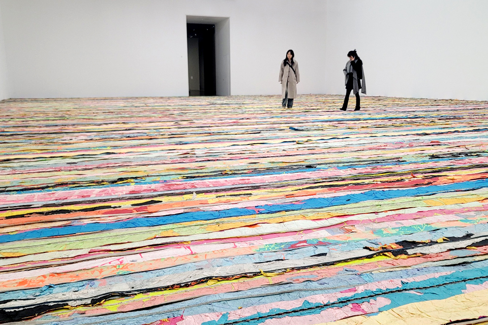 넓은 전시장 온 바닥에 상업 광고 전단지와 엔드 페이퍼가 형형색색의 띠지처럼 한쪽 끝에서 다른 쪽 끝까지 길게 이어지며 카펫처럼 설치되어 있다. 두 명의 관객이 그 위에 서서 작품을 내려다보고 있다. 