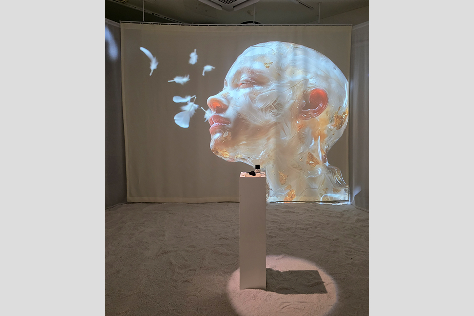 흰 모래가 깔린 공간에서 한 벽면을 꽉 채운 흰색 스크린에 영상이 투사되고 있다. 영상 속 반투명한 질감의 사람 얼굴에는 깃털이 덮여 있고, 얼굴 주변 한 방향으로 깃털이 날리고 있다.