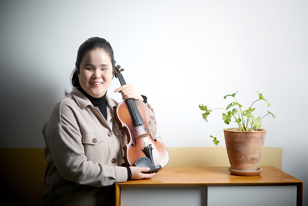 김지선 바이올리니스트가 작은 탁자에 기대어 서서 양손으로 바이올린을 몸에 가까이 잡고 포즈를 취하고 있다. 탁자 위에는 연두색 이파리의 식물 토분이 놓여 있다.