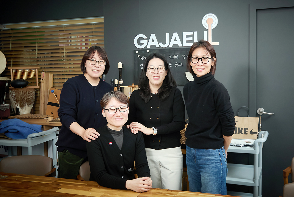 네 사람이 가까이 모여 밝게 미소지으며 카메라를 바라보고 있다. 이들 뒤로 짙은 회색으로 칠해진 벽 중앙에는 영문으로 ‘GAJAEUL’이라고 쓰여 있고, 벽을 따라 리코더, 차임벨 등 악기와 교구들이 놓여 있다.