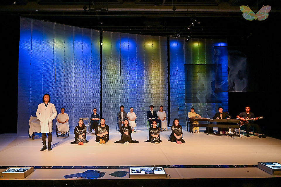 무대 앞줄에는 흰 가운을 한 배우가 서 있고, 옆으로 나란히 검은 의상을 입은 다섯 배우가 앉아 있다. 뒤편에는 혼자 혹은 들씩 짝지은 배우들과 키보드와 기타 등 라이브 연주자들이 의자에 나란히 앉아 있다. 뒷벽에는 천이 바닥까지 내려뜨려져 있고 파란색과 녹색 조명이 비추고 있다. 