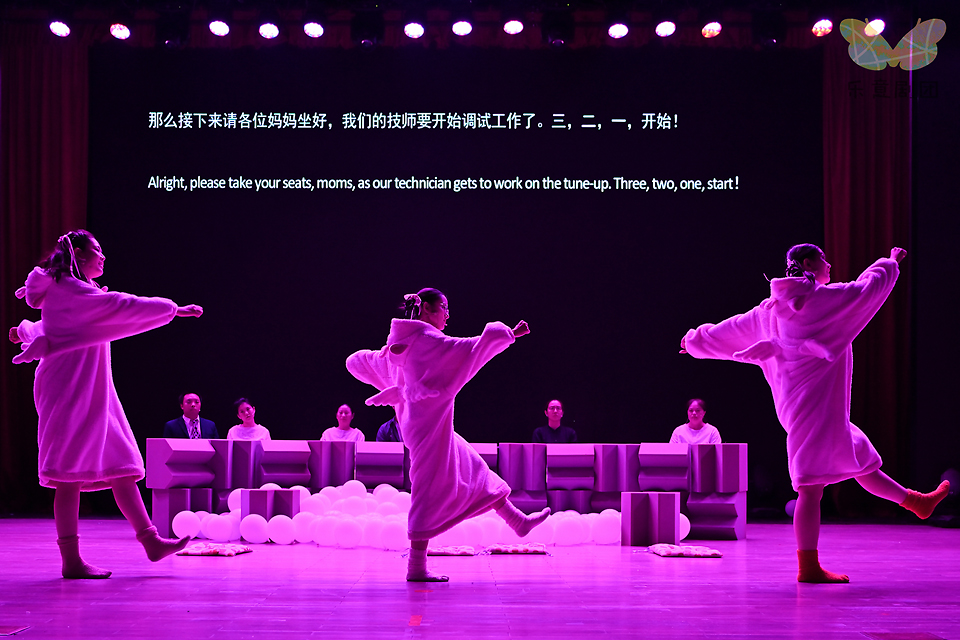 임부복 같은 흰 원피스를 입은 배우 세 명이 팔과 다리를 힘차게 들고 한 방향으로 걷고 있다. 뒤편으로 배우들이 나란히 책상을 두고 앉아 있고, 벽에는 중국어와 영어 자막이 영사되고 있다.