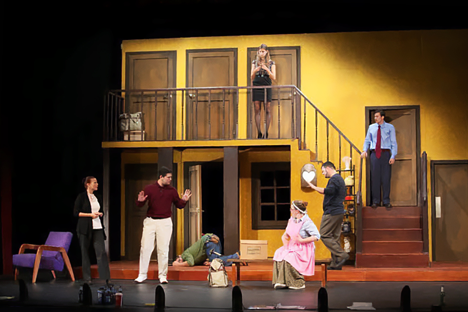 무대 위에 2층 구조의 현대 주택 세트가 설치되어 있고, 여러 명의 배우가 아래층과 계단, 위층 발코니 등에 흩어져서 아래층 문앞에 쓰러져 있는 한 사람을 바라보고 있다.