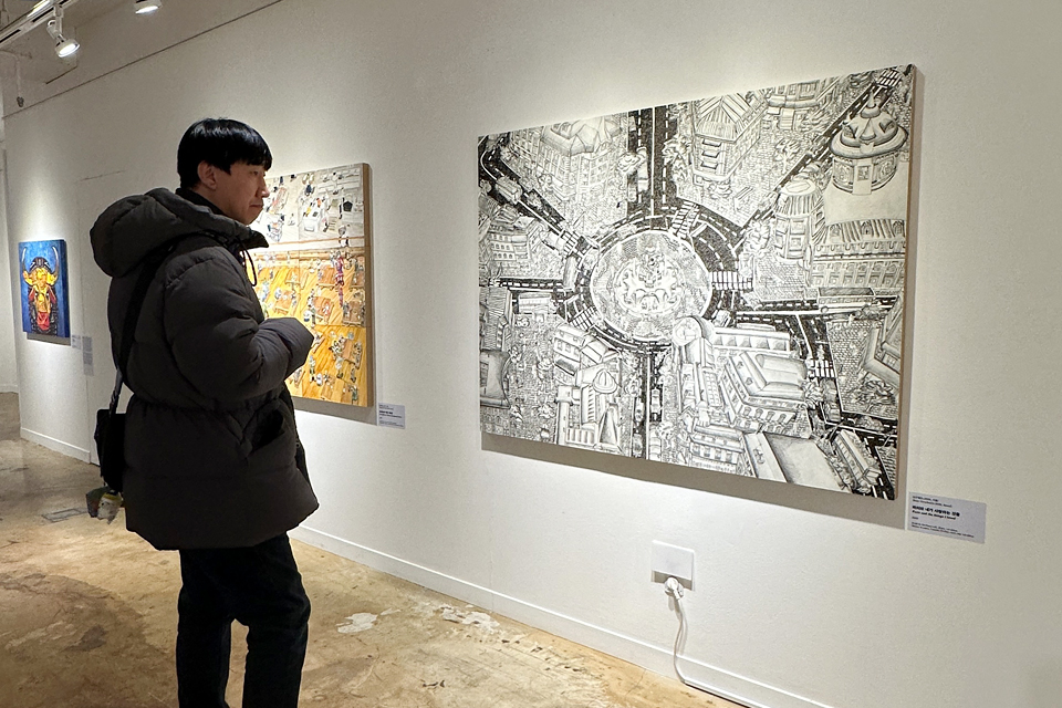 천장 트랙 조명 아래 여러 점의 회화가 나란히 걸려 있다. 양병철 활동가가 도시 풍경 드로잉 작품인 〈파리와 내가 사랑한 것들〉을 감상하고 있다.