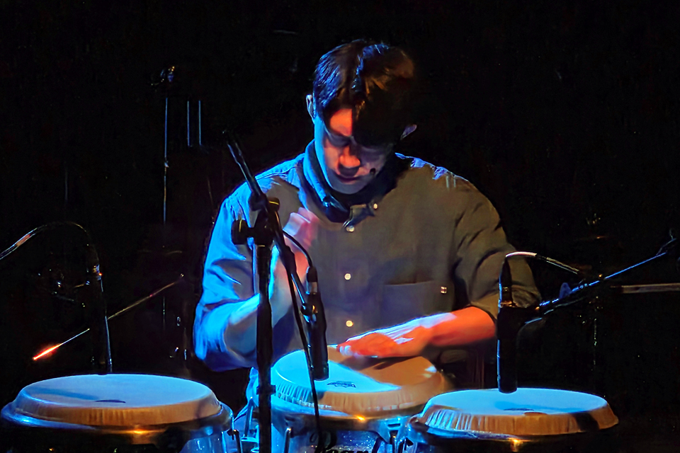 어두운 무대에서 파란색과 주황색 조명이 비치는 가운데, 이성재 퍼커셔니스트가 세 개의 콩가를 두 손으로 두드리며 연주하고 있다.