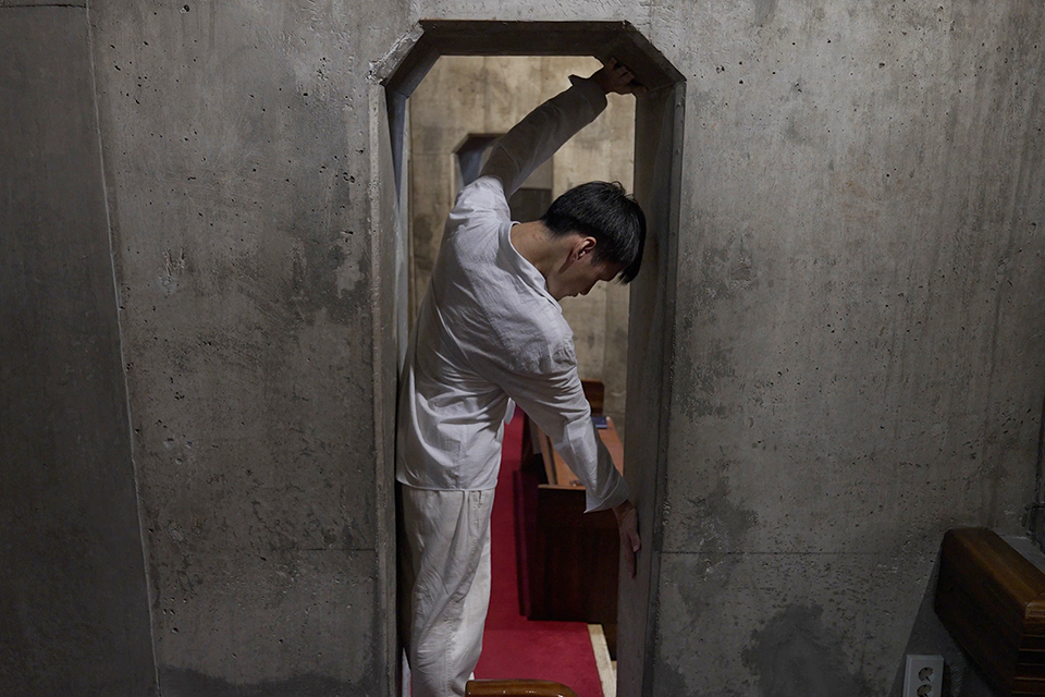콘크리트 벽으로 된 좁은 공간에서 흰 점프슈트를 입은 문승현 작가가 문틀에 몸을 기울이고 서서 팔과 다리로 문틀을 짚고 있다. 문 안쪽 바닥에는 붉은 천이 길게 깔려 있다.