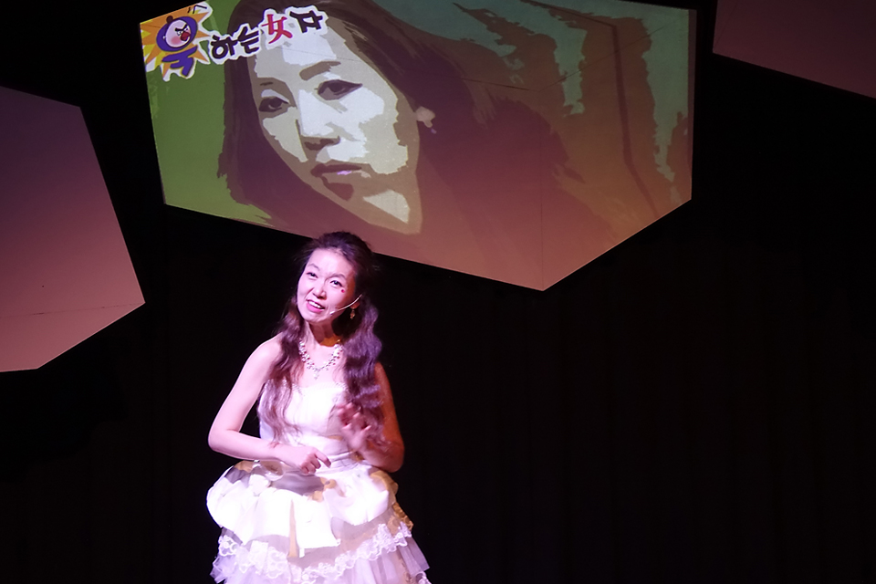 어두운 무대에서 흰색 드레스를 입고 긴 머리를 내려뜨린 박세영 배우가 이야기하고 있다. 뒤편 스크린 오브제에는 그래픽적으로 표현된 출연자의 얼굴이 투사되고 있고 “욱하는 女자”라고 쓰여 있다.