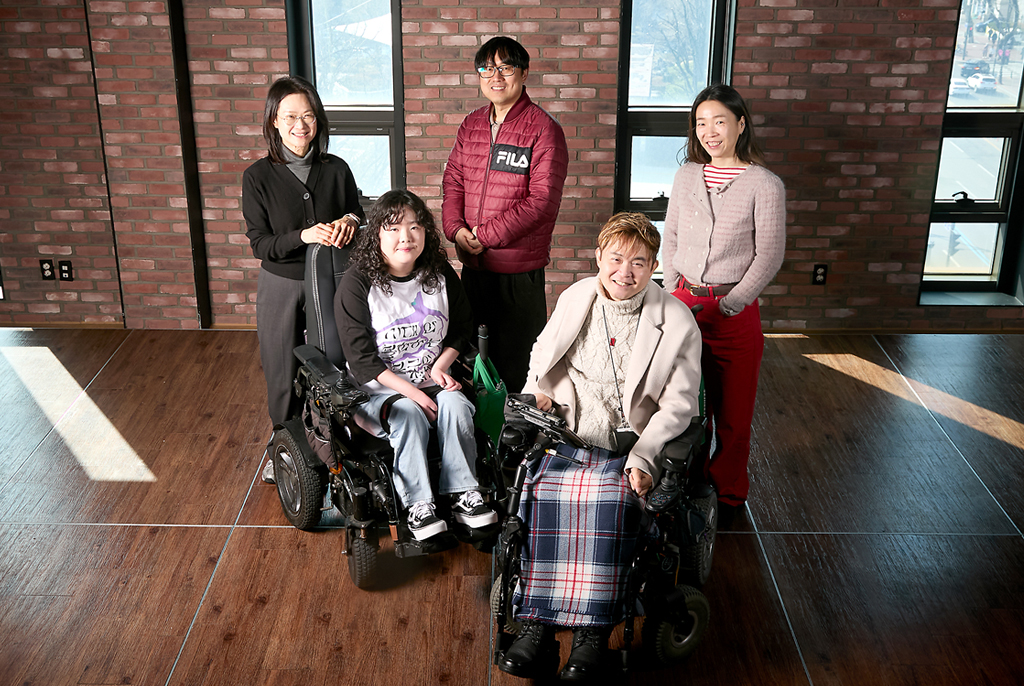 붉은 벽돌벽에 세로로 길게 난 창을 배경으로 다섯 명이 미소 지으며 카메라를 바라보고 있다. 앞의 두 사람은 휠체어에 앉아 있고, 뒤의 세 사람은 서 있다.