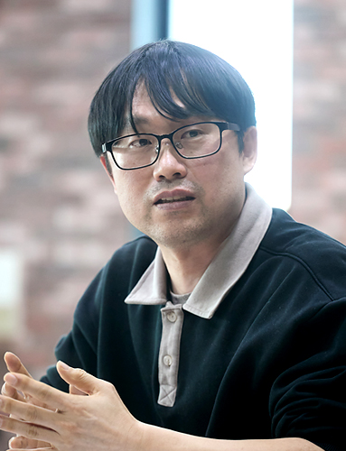 붉은 벽돌벽에 세로로 길게 난 창을 배경으로 다섯 명이 미소 지으며 카메라를 바라보고 있다. 앞의 두 사람은 휠체어에 앉아 있고, 뒤의 세 사람은 서 있다.