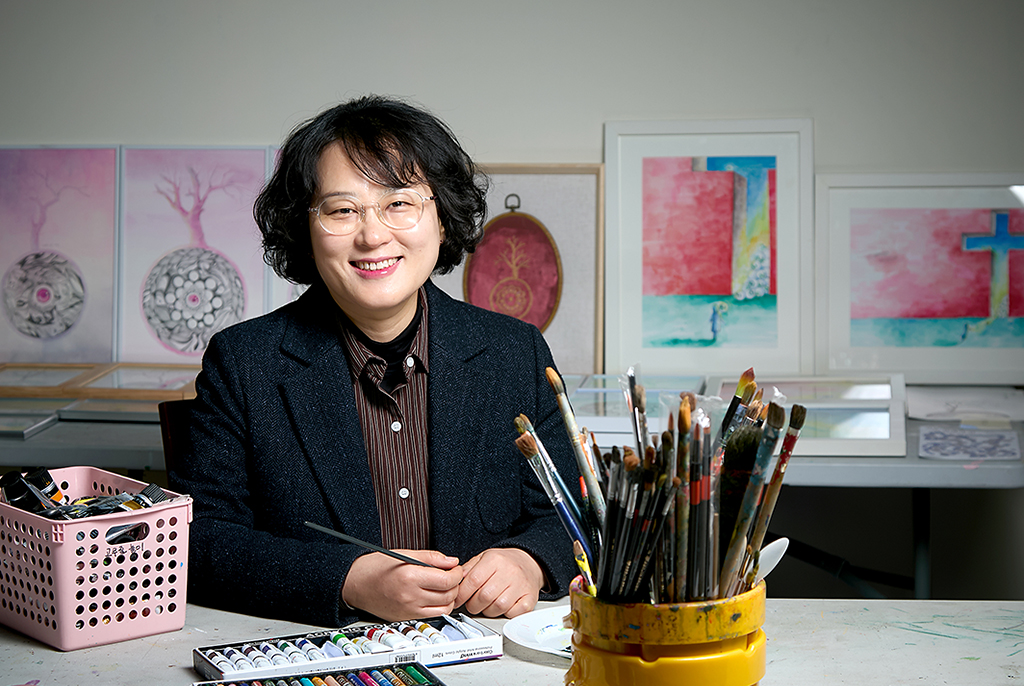 단발머리에 안경을 쓰고 검정 재킷을 입은 문귀화 작가가 테이블 앞에 포즈를 취하고 앉아 밝게 웃고 있다. 테이블 위에는 붓이 가득 꽂힌 통과 물감 상자, 오일파스텔 등이 놓여 있고, 뒤쪽 벽에 기댄 테이블 위에는 여러 점의 그림 액자가 놓여 있다.