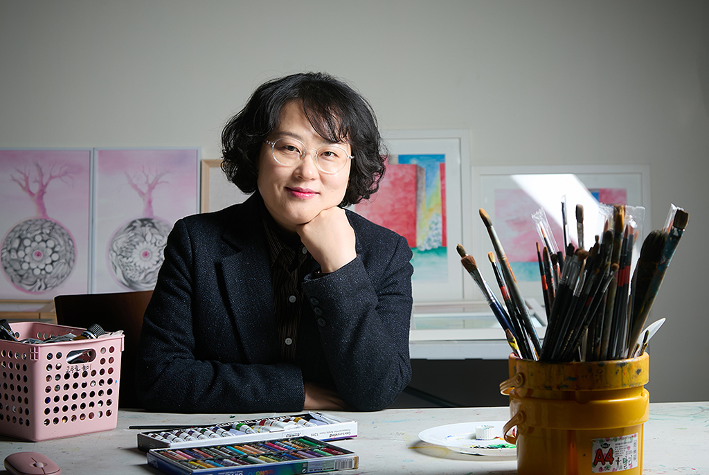 문귀화 작가가 테이블 위에 팔꿈치를 기댄 채 턱에 손을 괴고 앉아 살짝 미소 짓고 있다. 테이블 위에는 붓이 가득 꽂힌 통과 물감 상자, 오일파스텔 등이 놓여 있고, 뒤쪽 벽에 기댄 테이블 위에는 여러 점의 그림 액자가 놓여 있다.