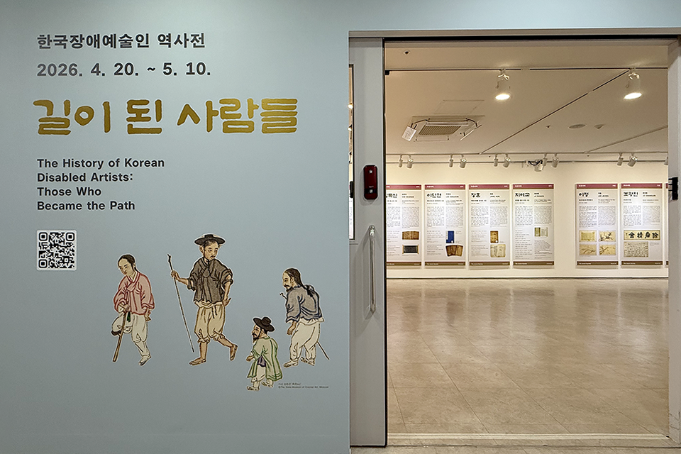 전시장의 입구와 내부가 함께 보이는 전경. 화면은 왼쪽의 전시 안내 벽면과 오른쪽의 열린 문틈으로 보이는 전시장 내부로 나뉜다. 오른쪽 벽면에는 “한국장애예술인 역사전” “길이 된 사람들” “2026.4.20.~5.10.”이라고 쓰여 있고 영문이 작게 쓰여 있고, 하단에 김준근의 풍속화 〈병신〉 작품 속 인물이 그려져 있다. 왼쪽 전시장 내부에는 벽을 따라 전시 패널이 나란히 걸려 있는 모습이 보인다.