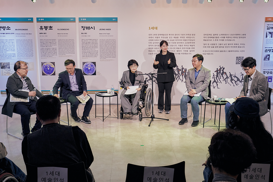 전시를 배경으로 다섯 사람이 의자에 앉아 이야기 나누고, 수어통역사 한 명이 통역하고 있다. 전경에는 경청하고 있는 관람객의 뒷모습이 보인다.