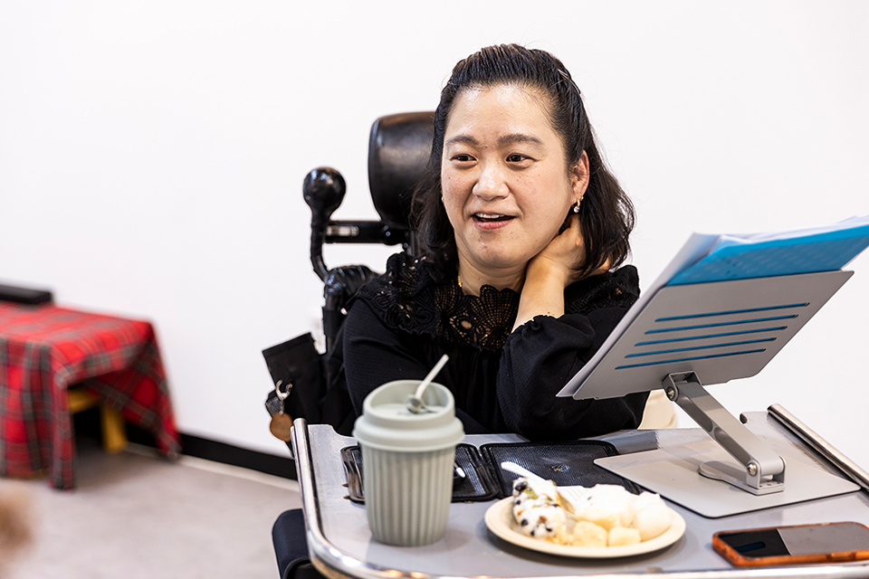 조우리 배우가 전동휠체어에 앉아 한 손을 턱 아래에 받친 채 옆을 바라보며 이야기하고 있다. 트레이 위에는 음료컵, 떡 접시, 스마트폰, 종이들이 올려진 태블릿 거치대가 놓여 있다.