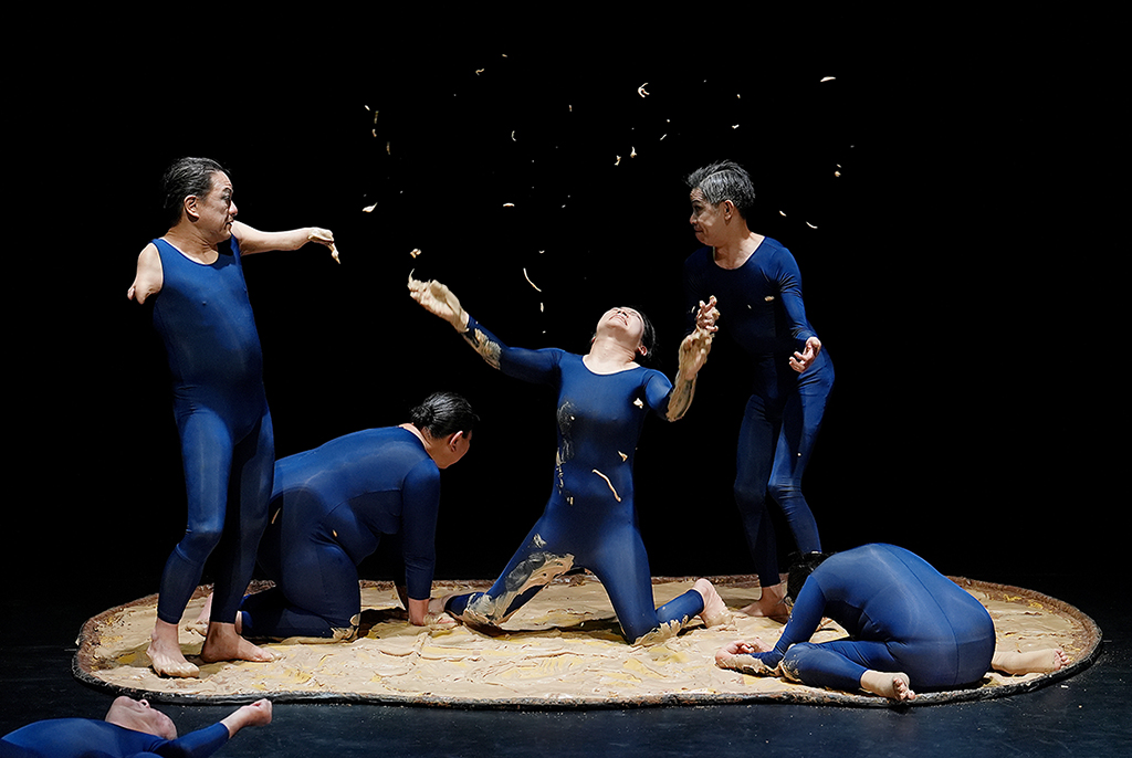 파란색 레오타드를 입은 공연자 다섯 명이 진흙이 깔린 원형 무대 위에 있다. 한 명은 무릎을 꿇고 두 팔을 위로 뻗어 머리를 젖히고 있으며, 나머지는 서 있거나 바닥에 엎드린 채 각자 다른 자세를 취하고 있다. 공연자들의 몸과 무대 바닥에 진흙이 묻어 있고, 공중에도 진흙 파편이 흩뿌려져 있다.