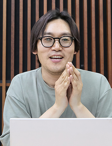 검정색 뿔테 안경을 쓰고 카키색 라운드 티셔츠를 입고, 두 손을 깍지 낀 채 턱 밑에 모은 남성이 노트북 앞에 앉아 정면을 바라보며 미소 짓고 있다. 