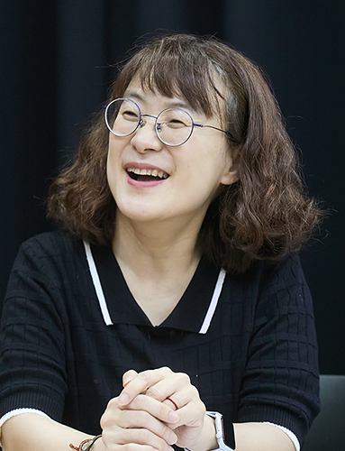 금속테 안경을 쓰고 단발머리 펌을 하고 검정색 티셔츠를 입은 여성이 두 손을 책상 위에 모은 채 옆을 보며 웃고 있다. 
