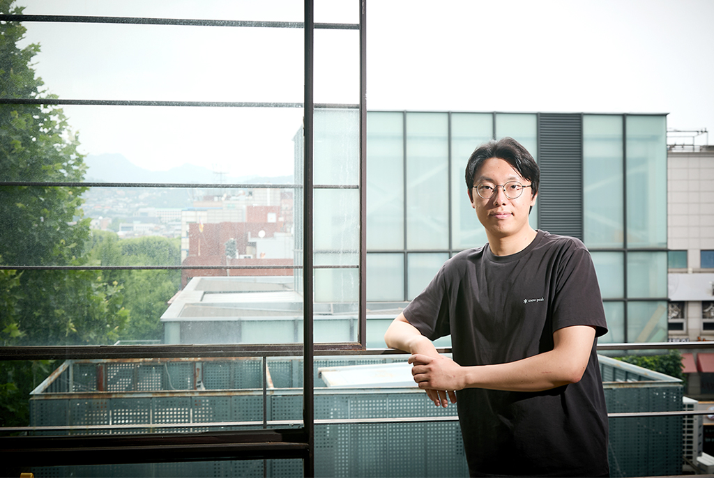 안경을 쓰고 검은색 반팔 티셔츠를 입은 작가가 한 팔을 철제 난간에 걸친 채 기대어 서서 정면을 바라보며 포즈를 취하고 있다. 뒤쪽으로는 가까이에 초록색이 짙은 큰 나무들과 건물 옥상, 멀리 낮은 건물이 빼곡한 도시 풍경이 펼쳐져 있다.