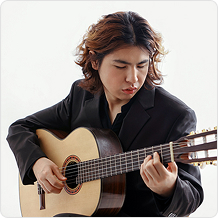 긴 곱슬머리에 정장을 입은 남성이 클래식 기타를 연주하는 포즈를 취한다.