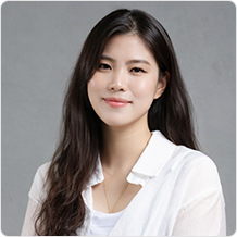 긴머리에 하얀색 셔츠를 입은 여성이 정면을 응시하고 있다.