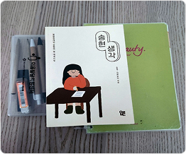 바닥에 볼펜, 연필, 지우개가 든 필통, 책 “송현 생각”, 연두색 노트가 겹쳐져 있다. 책에는 하얀 바탕에 책상 앞에 앉아 글을 쓰고 있는 소녀 삽화가 있고 “발달장애인 송현의 첫 에세이집” “송현, 아라보다 지음”이라고 쓰여 있다.
