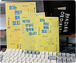 노트북 모니터와 컴퓨터 키보드 사이에 기대어 같은 책 세 권이 놓여 있다. 노란색 표지의 책에는 『여기는 안녕시 행복동입니다』라고 쓰여 있다.