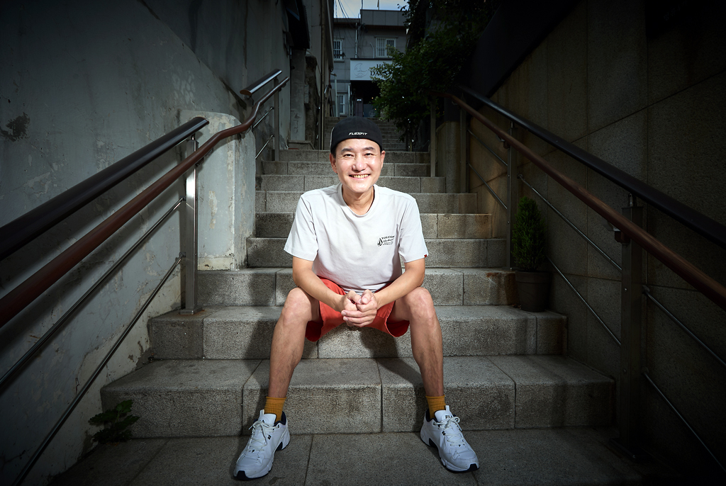 이광석 무용가가 가파른 골목길 돌계단 중앙에 앉아 있다. 모자를 뒤로 눌러쓰고 흰색 반팔 티셔츠와 주황색 반바지, 흰색 운동화를 착용하고 있다. 