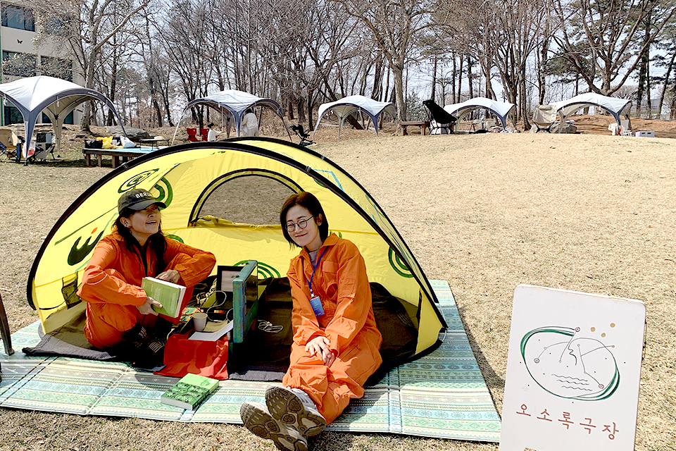 야외 잔디밭 위, 매트가 깔린 노란색 소형 팝업 텐트 안에 주황색 점프수트를 입은 두 사람이 앉아 있다. 텐트 앞에는 ‘오소록극장’이라는 글씨와 그림이 그려진 나무 입간판이 놓여 있고, 뒤쪽으로는 숲을 배경으로 여러 개의 돔 텐트가 설치되어 있다