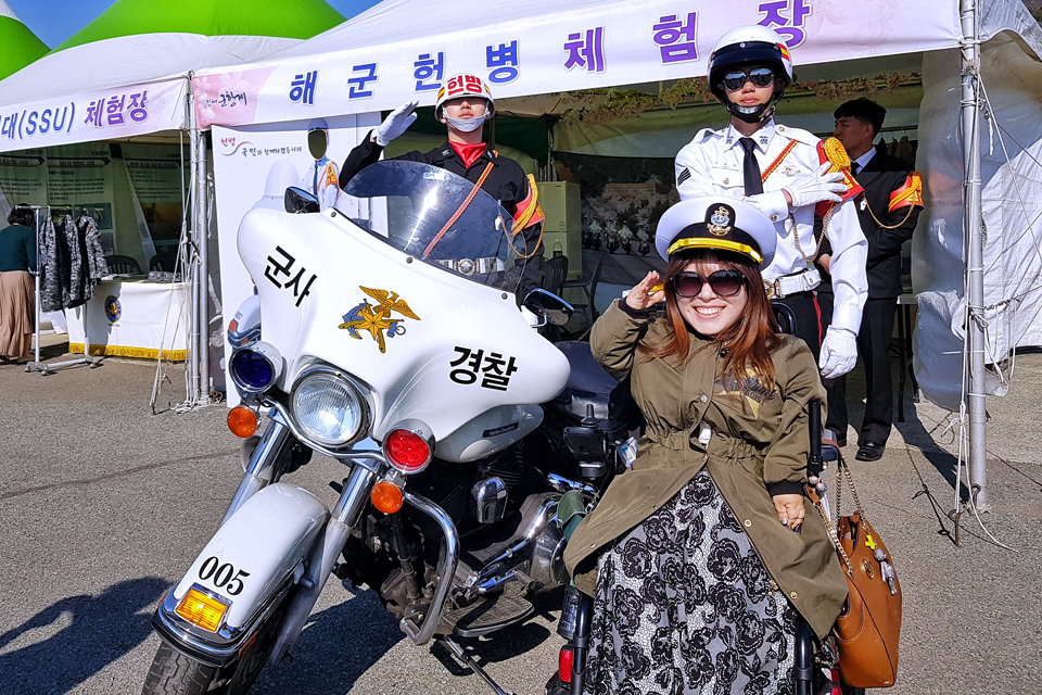 ‘해군헌병체험장’ 현수막이 걸린 천막 앞에서 필자가 해군 모자를 쓰고 전동휠체어에 앉아 경례 포즈를 하며 웃고 있다. 그 뒤로 해군 정복과 헌병 제복을 입은 대원 두 명이 흰색 군사경찰 오토바이 옆에서 경례 포즈를 취하고 있다.