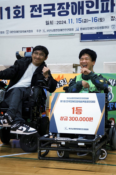 대회 현수막 아래에서 전동휠체어를 탄 홍성훈이 “1등 상금 300,000원”이라 적힌 패널을 들고 환하게 웃고 있다.