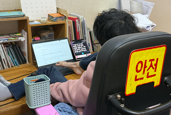 휠체어에 앉아 오른발 발가락으로 책상 위에 놓인 노트북으로 타이핑하고 있는 필자의 뒷모습. 노트북 옆에는 패드에 큰 글씨가 띄워져 있다. 책상 위에는 자료와 문구가 가지런히 정돈되어 있다.
