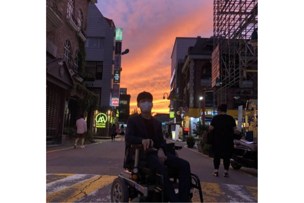 해질녘 붉은 석양으로 물든 하늘과 대학로 거리를 배경으로 전동휠체어를 탄 필자가 정면을 응시하고 있다.