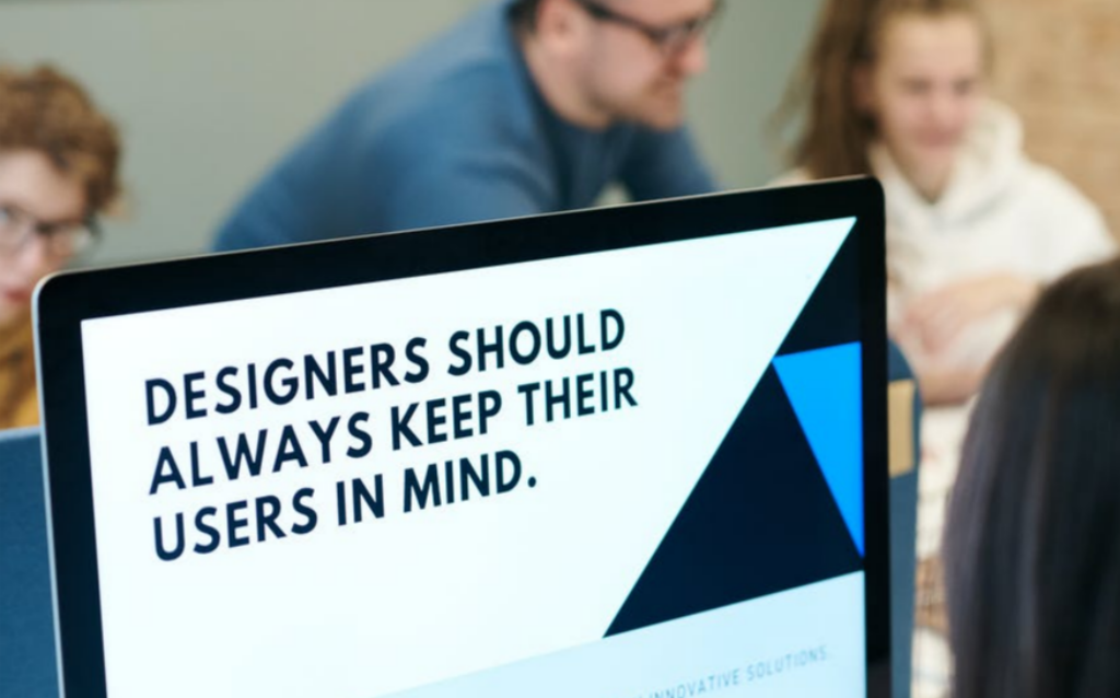 컴퓨터가 있는 사무실 내부, 사람들이 함께 일하고 있다. 컴퓨터 모니터에는 DESIGNERS SHOULD ALWAYS KEEP THEIR USERS IN MIND(설계자는 항상 사용자를 염두에 두어야 한다.)라는 문장이 적혀있다.